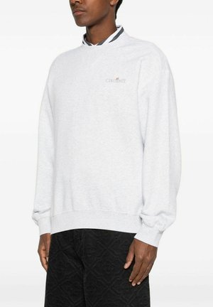 Mand iført lysegrå Carhartt sweatshirt og sorte mønstrede bukser, stående mod hvid baggrund, vendt let mod venstre.