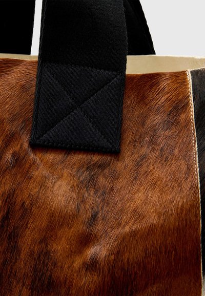 Gros plan d'un sac marron à texture de fourrure avec des poignées en tissu noir épais cousues en grand motif en X aux points de fixation.