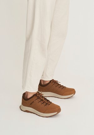 Bruine veter sneakers met dikke witte zolen gedragen bij crèmekleurige broek op een effen lichte achtergrond.