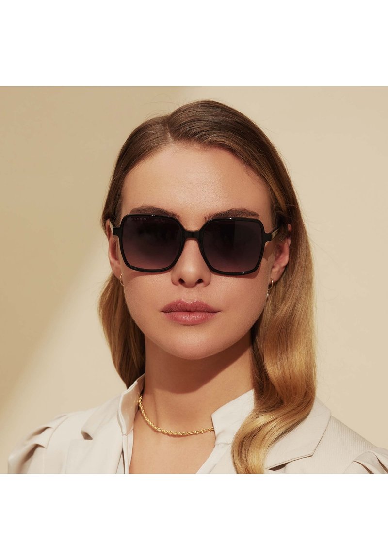 Lunettes de soleil carrées avec des verres dégradés foncés et une monture noire. Le modèle porte une chemise couleur crème et un collier en chaîne dorée.