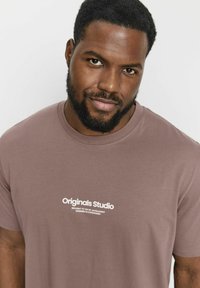 Brun bomuld t-shirt med rund halsudskæring og hvidt logo-tryk, der står "Originals Studio" på forsiden.