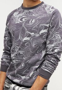 Man in een sweatshirt met lange mouwen, voorzien van een grijs en wit marmerpatroon, gecombineerd met een ruitjesbroek en een casual uitstraling.