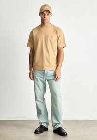 Ταν βαμβακερό μπλουζάκι με τσέπη στο στήθος, ανοιχτό γαλάζιο τζιν και μαύρα παπούτσια. Το καπέλο είναι ταν με το λογότυπο "BOSS" σε λευκό. Απλή, χαλαρή εφαρμογή.