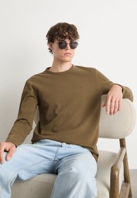 Olívazöld bordázott, hosszú ujjú póló, kerek nyakkivágással, világoskék farmernadrággal párosítva. A modellen kerek fekete napszemüveg van, ülő pozícióban.