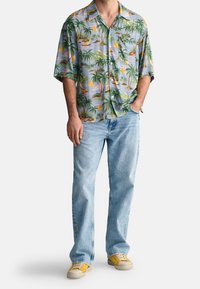 Muž stojící s jednou rukou v kapse, oblečený v krátkém tričku s tropickým potiskem, světle modrých džínách a žlutých teniskách.