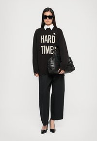 Black sweater with "HARD TIMES" in light beige, paired with wide black trousers. Black structured handbag, pointed black heels, and sunglasses.