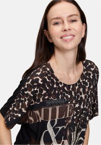 Brązowa koszulka w panterkę z krótkim rękawem, okrągłym dekoltem i metalicznymi akcentami z napisem "UŚMIECH" w wyraźnym druku.