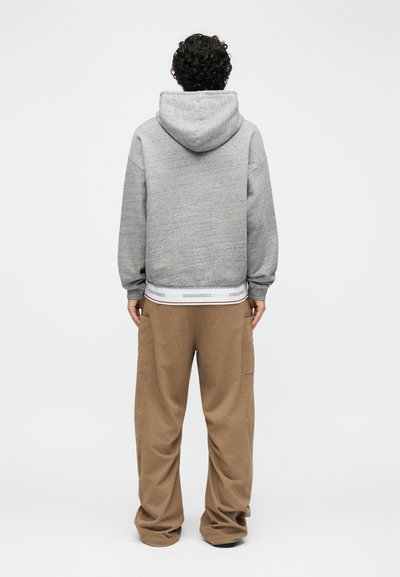 Persona con capelli ricci vista di spalle, indossa una felpa con cappuccio grigia e pantaloni marroni larghi con tasche laterali su sfondo neutro.