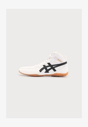 Baskets montantes blanches Asics avec des bandes noires du logo et une semelle marron clair sur un fond uni clair.