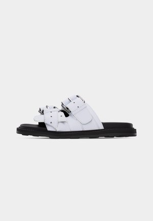 White leather sandal with two adjustable buckle straps and a black flat sole, shown from the side on a plain background.