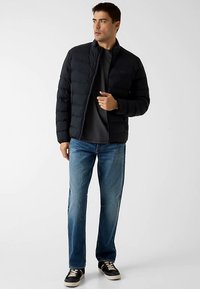 Schwarze Steppjacke mit horizontalen Nähten, hohem Kragen und Frontreißverschluss, kombiniert mit blauer Jeans und schwarzen Turnschuhen vor neutralem Hintergrund.