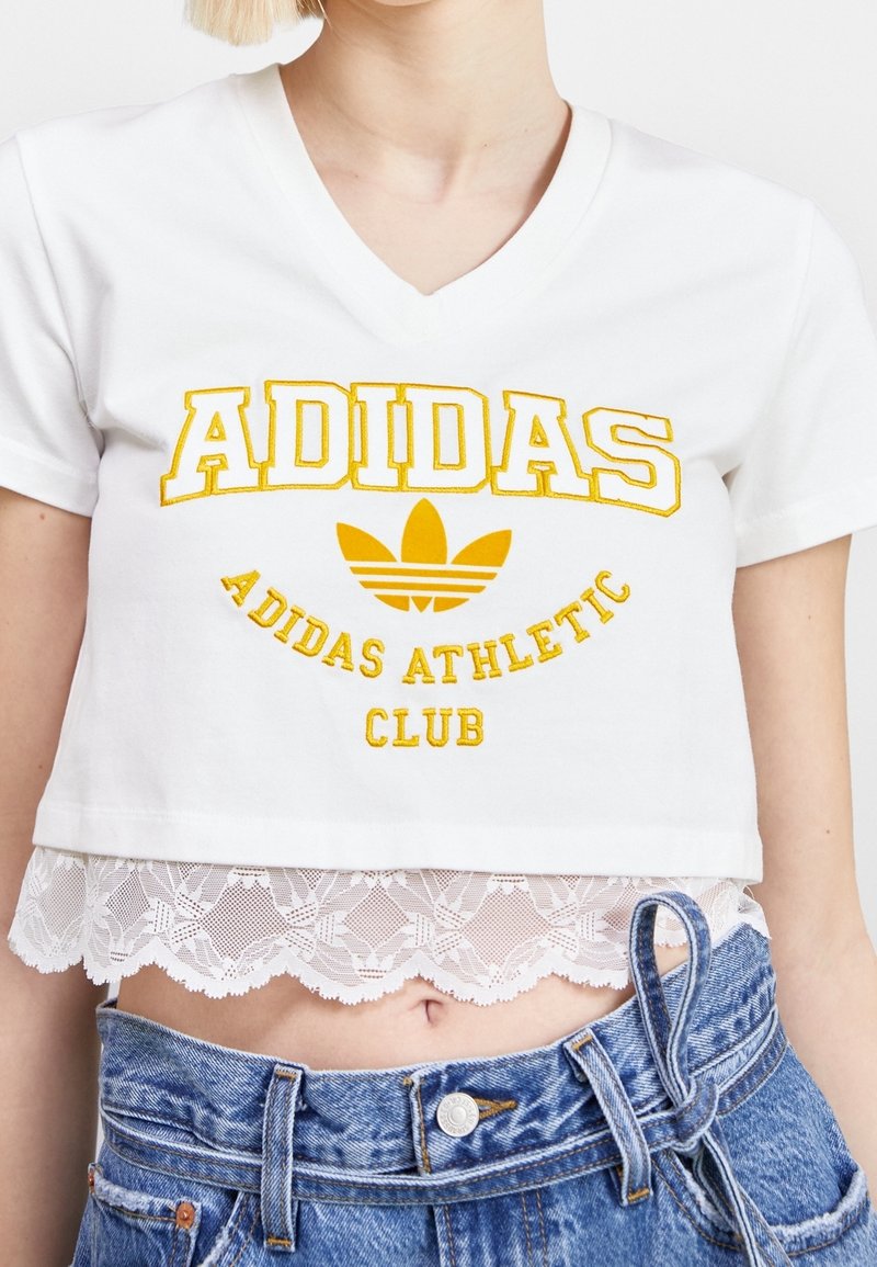Vit kortärmad t-shirt med gul broderad "ADIDAS" logotyp och "ADIDAS ATHLETIC CLUB", med spetskant i nederkanten.