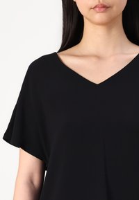 Blusa negra de cuello en V con mangas cortas, hecha de tejido suave, presenta un diseño simple y un corte holgado, enfatizando el minimalismo.