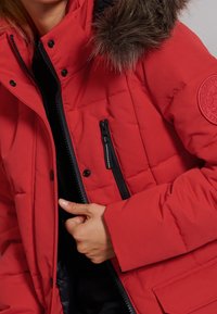 Rode gewatteerde jas met een afneembare capuchon van bont, een ritszak en drukknoopsluiting aan de voorkant. Soepele textuur met een doorgestikt ontwerp.