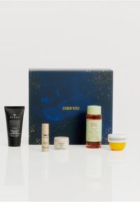 En uppsättning av fem hudvårdsprodukter på en reflekterande vit yta. Inkluderar ett svart rör, tre små burkar och en grön flaska, arrangerade i en låda med navyblå och guld design.