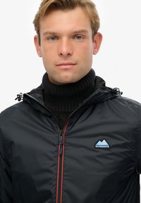 Sort letvægtsjakke med hætte, featuring en rød lynlås og et blåt bjerglogo. Båret over en sort ribbet turtleneck sweater.