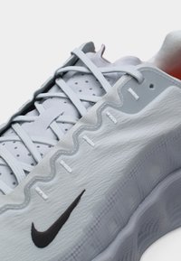 Κοντινή λήψη ενός λευκού και γκρι αθλητικού παπουτσιού Nike με μαύρο λογότυπο swoosh, λεπτομερείς ραφές και κορδόνια σταυρωτά σε υφασμένη σόλα.