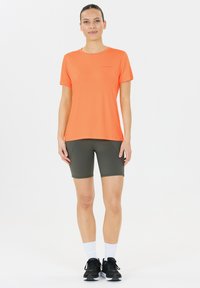 Kirkkaan oranssi urheilupaita lyhyillä hihoilla, yhdistettynä oliivinvihreisiin shortseihin. Mustat lenkkarit ja valkoiset sukat täydentävät asukokonaisuuden.