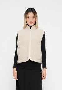 Žena v béžovej bezrukávovej fleecovej veste na zips, vrstevnej na čiernu dlhú blúzku a čiernu sukňu, stojí na bielom pozadí.