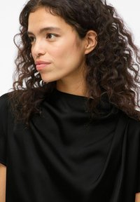 Top in satin nero con scollatura drappeggiata e maniche corte. Il tessuto presenta una texture liscia, evidenziando morbide pieghe e contorni.