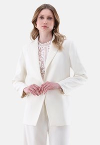 Weißer taillierter Blazer mit glatter Textur, Reverskragen und kontrastierendem gemustertem Innenfutter, kombiniert mit einem Spitzenoberteil.