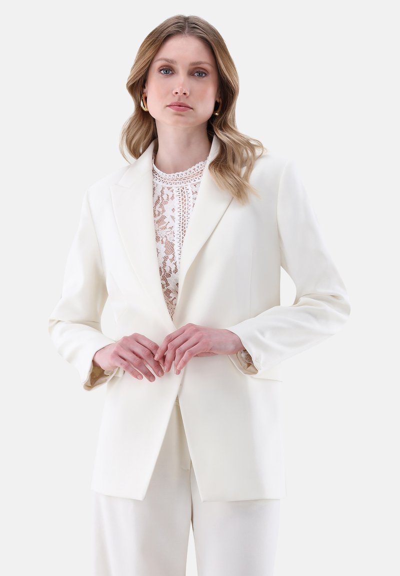 Weißer taillierter Blazer mit glatter Textur, Reverskragen und kontrastierendem gemustertem Innenfutter, kombiniert mit einem Spitzenoberteil.