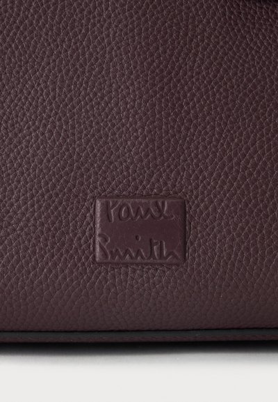 Surface en cuir texturé marron foncé avec un patch cousu embossé du nom de la marque "Paul Smith" en écriture cursive.