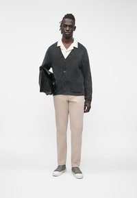 Graue gerippte Strickjacke mit V-Ausschnitt, Vorder Taschen und Knopfverschluss, kombiniert mit cremefarbenem Hemd und beiger Hose, hält eine schwarze Tasche.