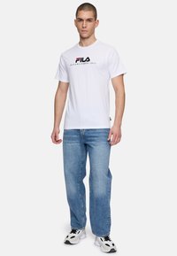 Camiseta de manga corta blanca con "FILA" impreso en negro y rojo, combinada con jeans de mezclilla azul claro y zapatillas negras y blancas.