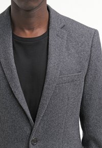 Grå ullblazer med en notch lapel, enkel knappstängning och sidofickor. Bärs över en svart T-shirt som visar ett texturerat tyg.