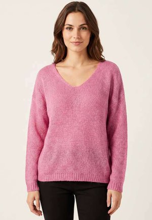 Femme portant un pull rose à manches longues et col en V et un pantalon noir, debout devant un fond clair uni, regardant vers l'avant.