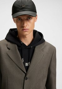 Gray textured blazer over a black hoodie with a logo. The cap is black with a shiny emblem at the front, blending casual and formal styles.