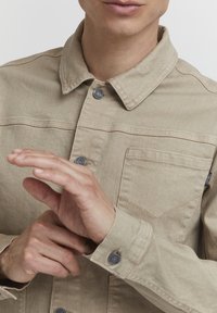 Beige denim jacket with a collared neck, front pocket, and silver snap buttons. The fabric has a smooth texture and visible stitching details.