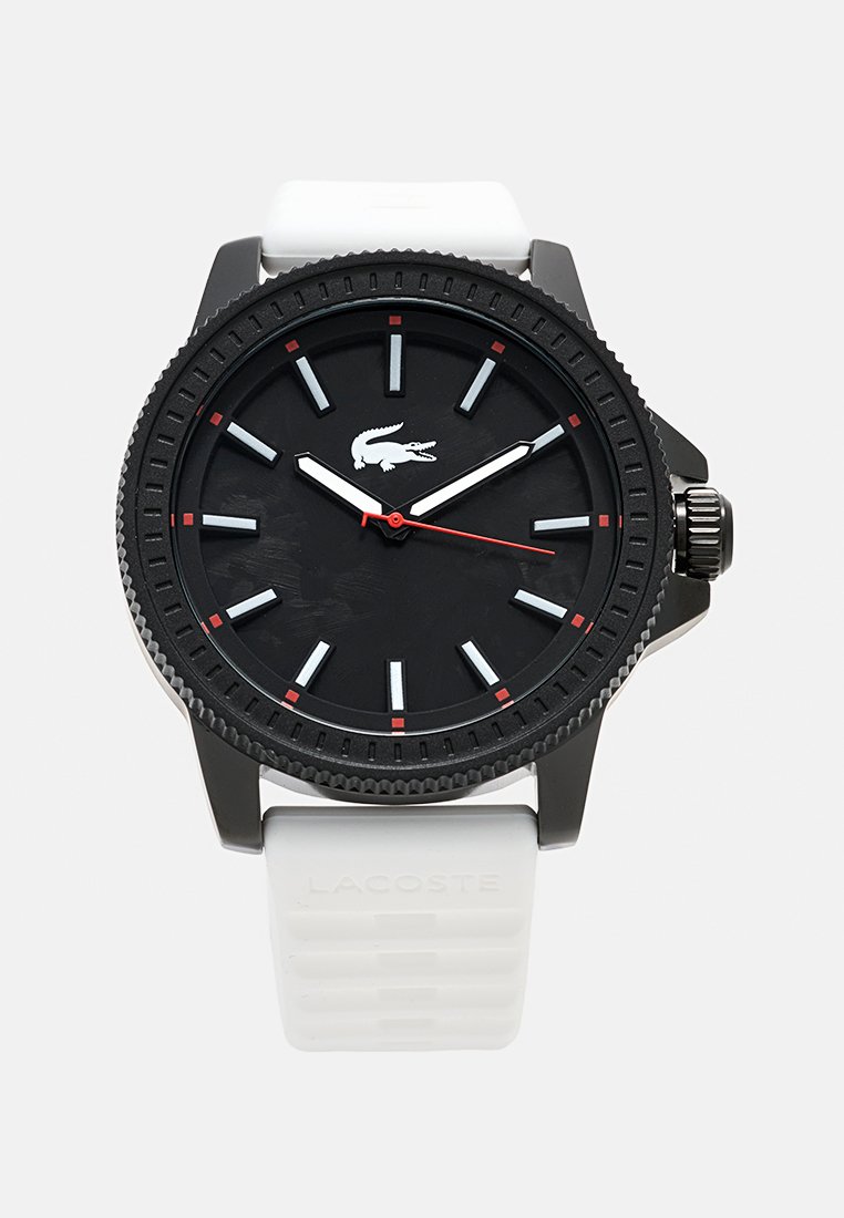 Montre analogique noire avec un bracelet blanc en plastique, présentant un cadran noir texturé, des accents rouges et des marqueurs d'heure en argent. Logo Lacoste affiché.