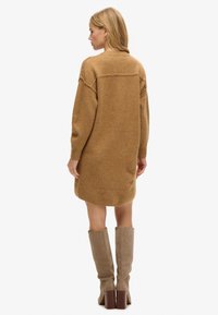 Vestido de punto en color marrón claro, con un corte holgado, hombros caídos y puños acanalados. Combinado con botas taupe hasta la rodilla que tienen un tacón cuadrado.