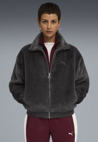 Person in gray plush zip-up jacket and maroon Puma tracksuit pants with white stripe, standing against a gray gradient background.