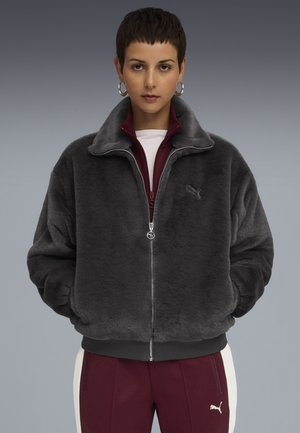 Person in gray plush zip-up jacket and maroon Puma tracksuit pants with white stripe, standing against a gray gradient background.