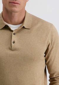 Beige Poloshirt aus einem weichen, strukturierten Stoff, mit drei schwarzen Knöpfen und einem klassischen Kragen-Design. Über einem weißen Unterhemd getragen.