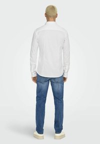 White long-sleeve shirt with a collar, displayed from the back. Paired with blue denim jeans and light-colored shoes. Smooth texture.