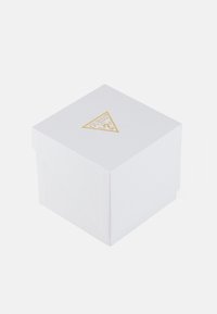 Hvit firkantet gaveeske med teksturert overflate, som har en gulltriangulær GUESS-logo på toppen. Lokk som er litt åpent.