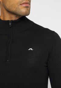 Black zip-up sweater with ribbed collar, featuring a small white logo on the chest. Material appears smooth and fitted.