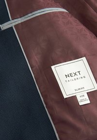 Bordeaux en marine jas interieur met geometrische textuur, voorzien van een wit label met de tekst "NEXT TAILORING SLIM FIT" en blauwe piping details.