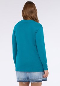 Niebieska dzianinowa bluzka z długim rękawem, z okrągłym dekoltem, żebrowanym wykończeniem i guzikami na mankietach. Miękki materiał o gładkiej teksturze.