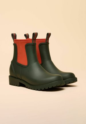 Grüne Gummistiefeletten mit roten elastischen Seiteneinsätzen, strukturierter schwarzer Sohle und Zuglaschen. Glatte Oberfläche mit mattem Finish.