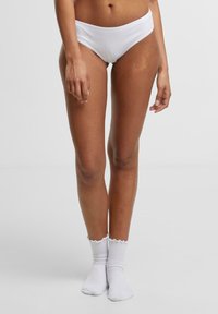 Bijeli pamukasti bikinij gaćice s niskim strukom. Model također nosi bijele rebraste čarape s valovitim rubom. Glatka tekstura, uski oblik.