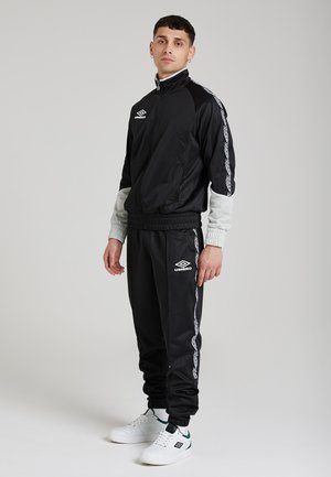 Tuta nera con accenti grigi, caratterizzata da un colletto alto, zip frontale e logo. Pantaloni affusolati con polsini elasticizzati e strisce laterali a motivi. Sneaker bianche.