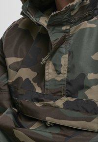 Gros plan d'une veste camouflage verte, marron et noire avec un col zippé et des cordons de serrage.