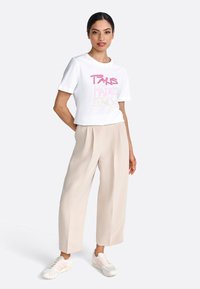 Tricou alb cu mânecă scurtă, cu text roz și auriu "PARIS MADRID", care are o croială lejeră. Asociat cu pantaloni săi largi bej și adidași.