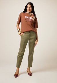 Camicia marrone con grafica "VINTAGE", pantaloni verde oliva e décolleté a punta in vernice bordeaux. Accessoriato con una sciarpa beige e bracciali dorati.