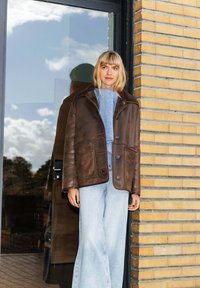 Veste en cuir brun avec un col en mouton, trois boutons à l'avant et une coupe décontractée, assortie à un jean large bleu clair.